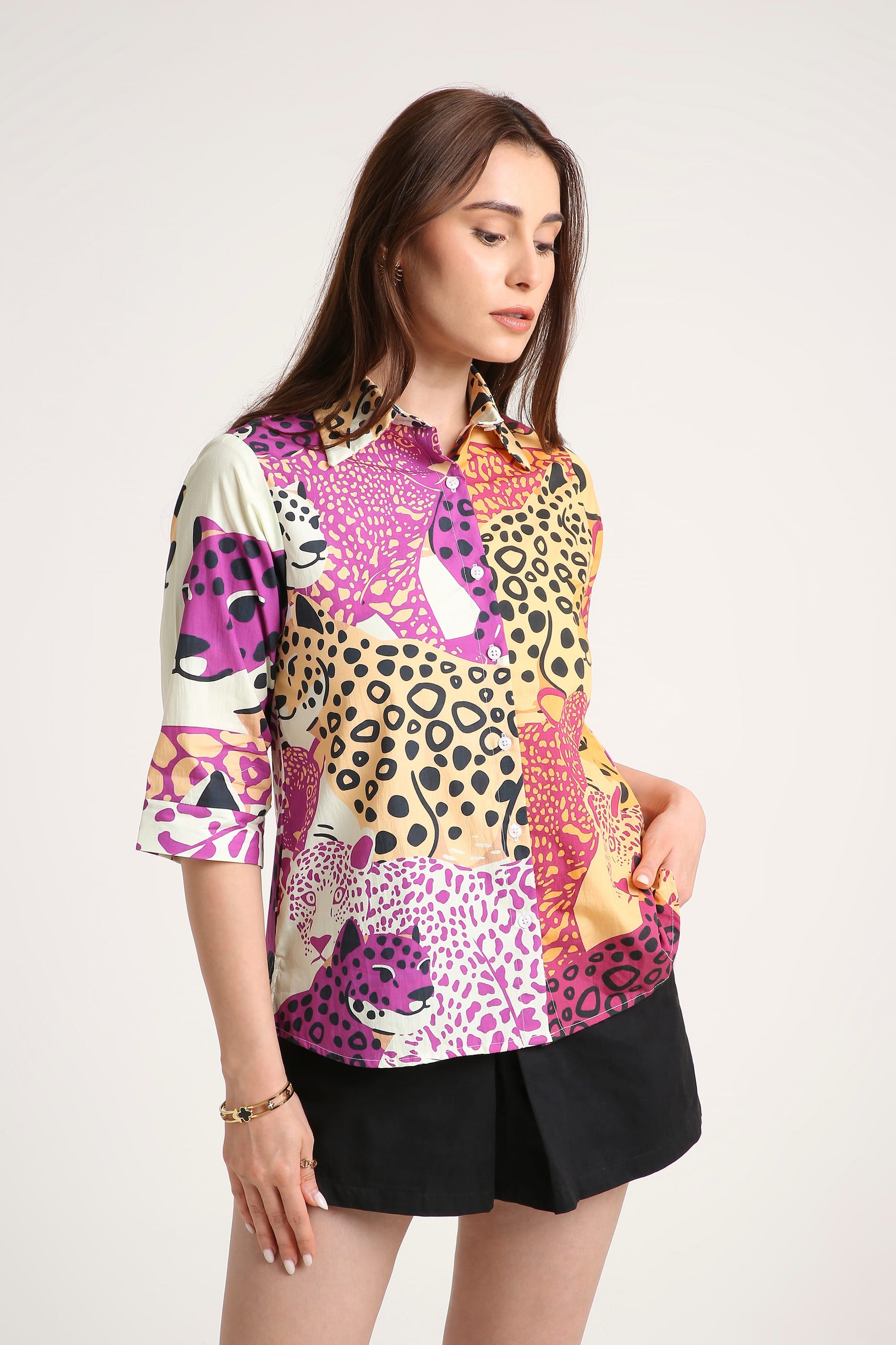 A woman wearing a short-sleeve button-up shirt with a vibrant multicolour animal print pattern featuring pink, purple, black, and beige leopard and cheetah motifs, paired with black shorts against a plain light background.