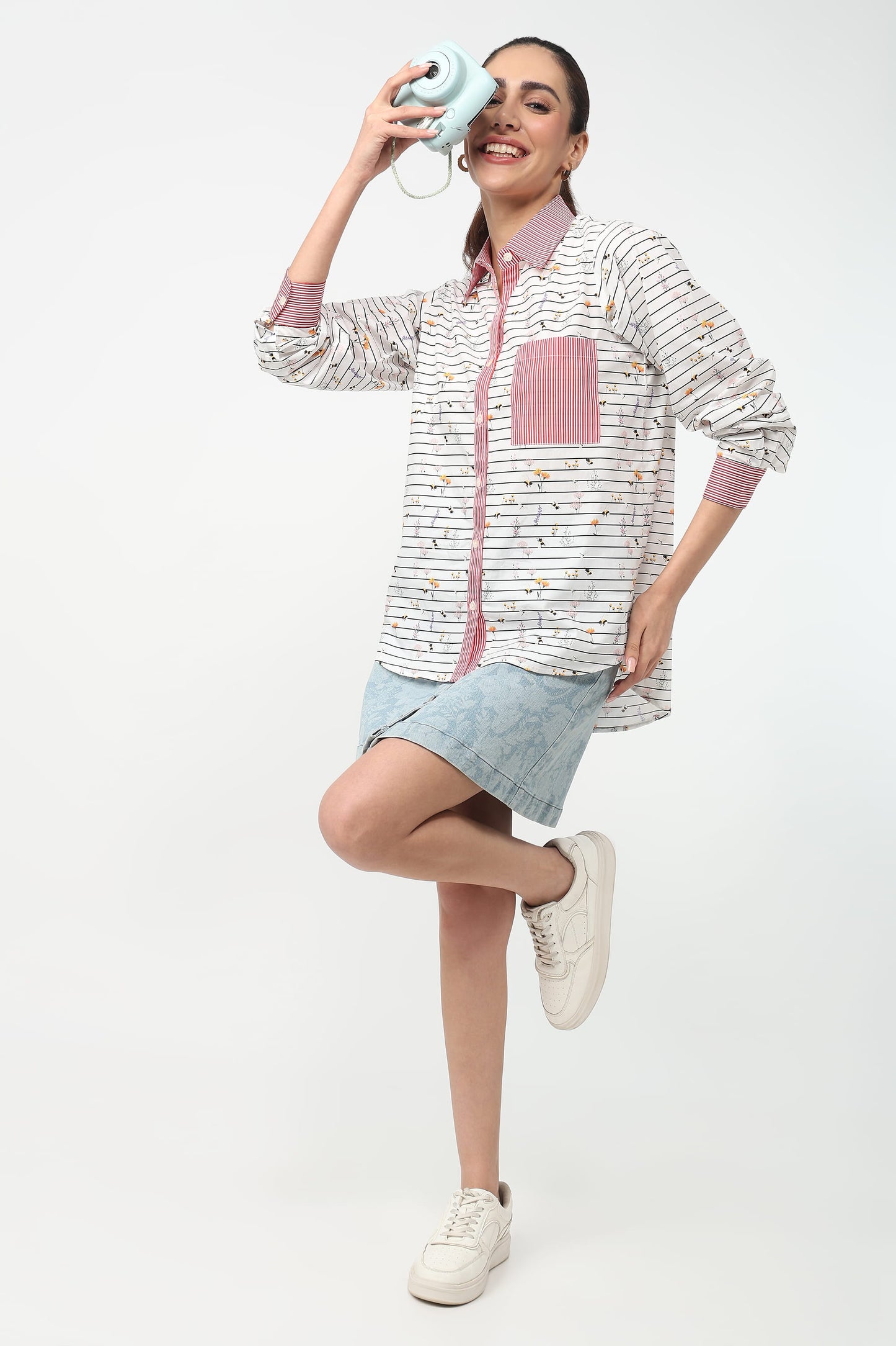 A woman modeling a long-sleeve button-up shirt with a white and gray striped pattern, a pink pocket, and a pink collar, paired with a denim skirt and white sneakers, against a plain white background.