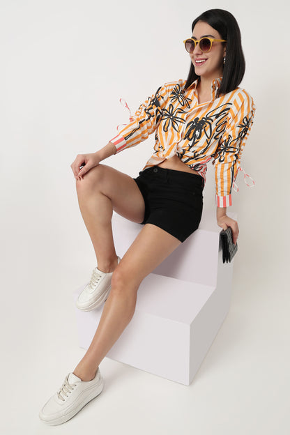 Woman wearing a patterned top and black shorts sitting on a white block against a white background