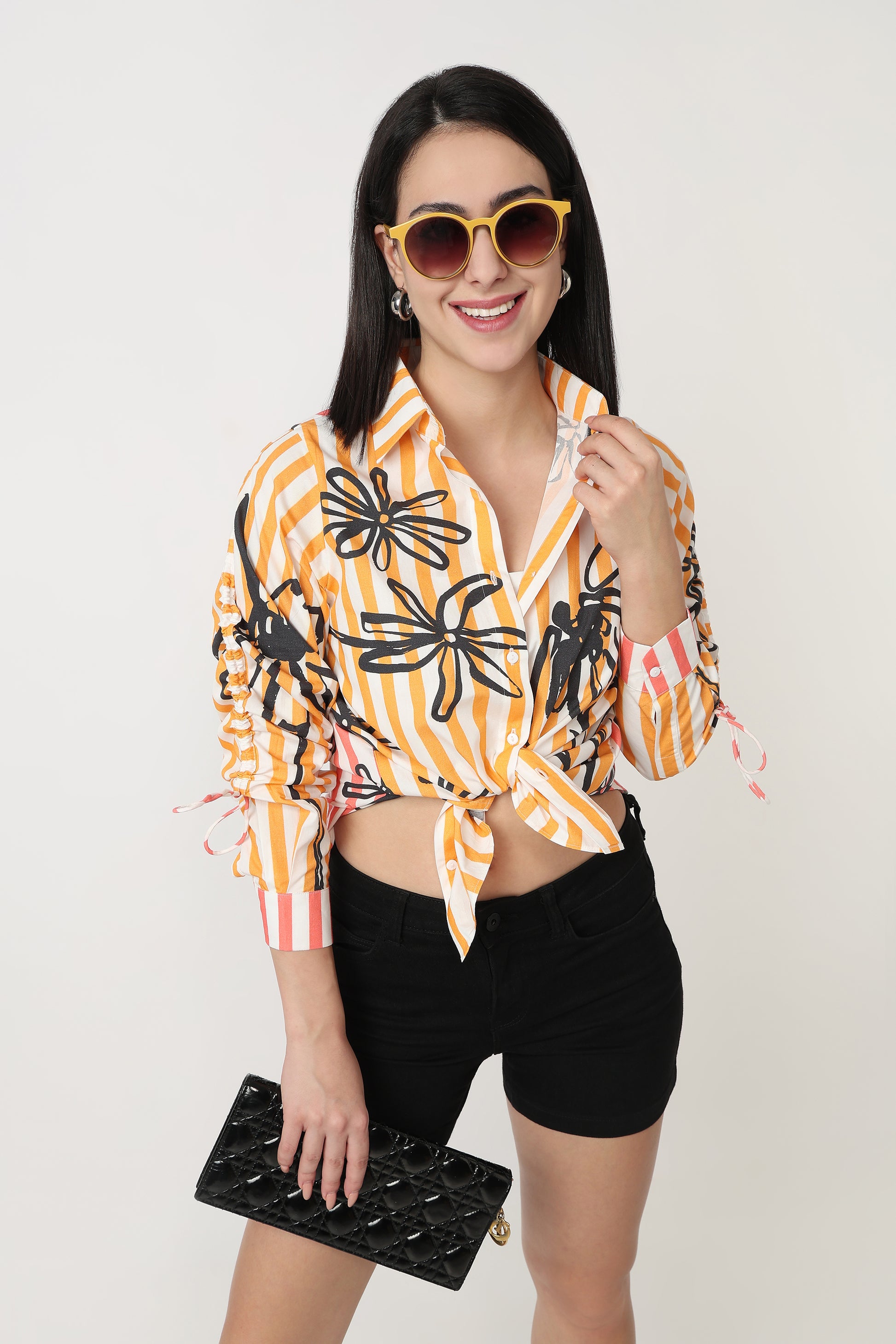Woman wearing a colorful striped shirt with floral patterns, black shorts, and sunglasses, holding a black clutch.