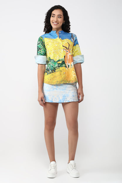 A woman modeling a matching short-sleeve button-up shirt and mini skirt set with a vibrant, multicolor landscape print featuring a deer, on a light gray studio background.