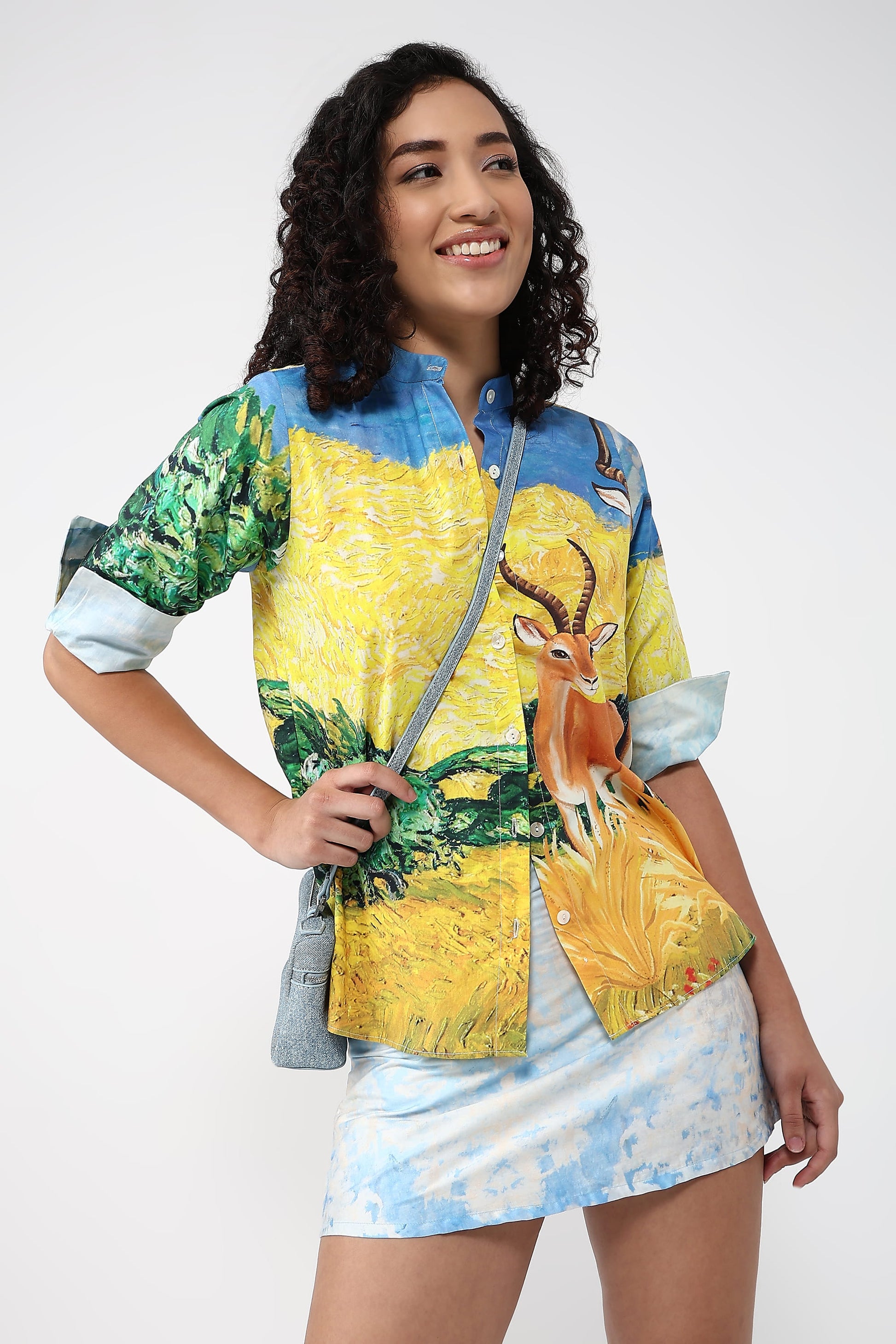 A woman modeling a short-sleeve button-up shirt with a vibrant, artistic print featuring a yellow sunflower field, green foliage, and a deer, paired with a light blue and white tie-dye mini skirt against a plain white studio background.