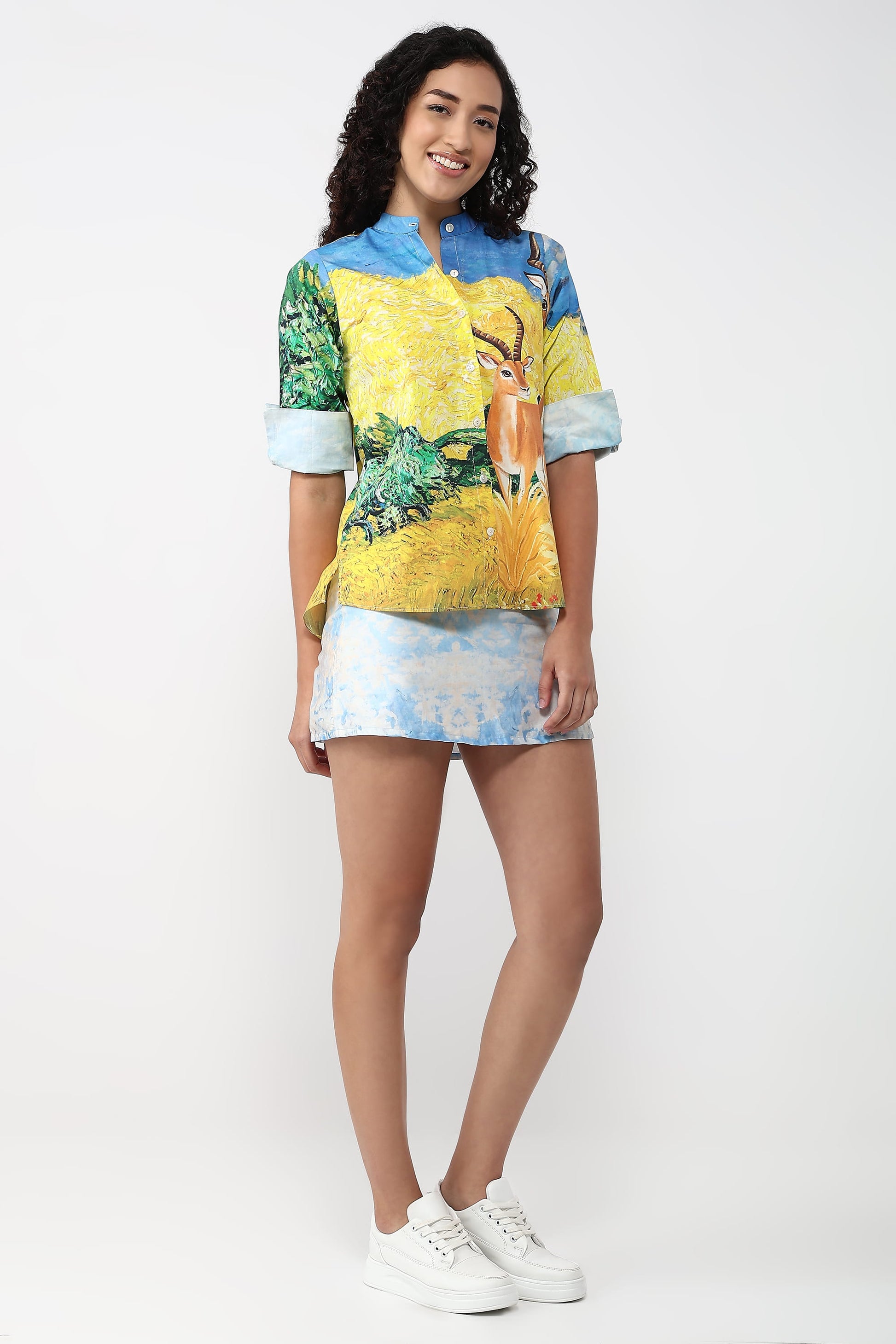 A woman modeling a short-sleeve button-up shirt with a colorful nature print featuring a deer and blue sky, paired with a matching mini skirt and white sneakers, against a plain white studio background.