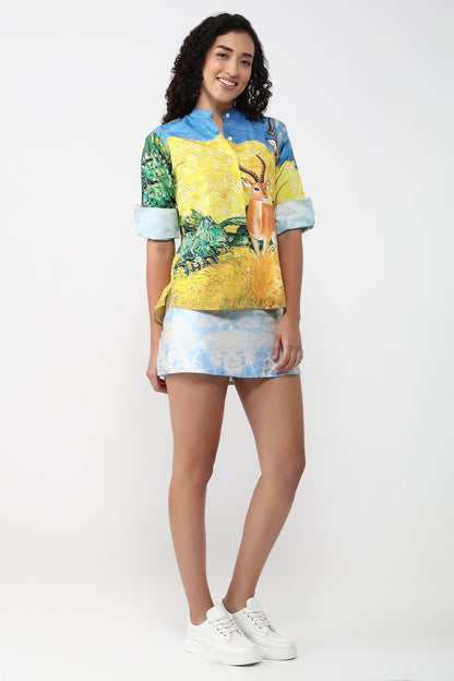 A woman modeling a short-sleeve button-up shirt with a colorful nature print featuring a deer and blue sky, paired with a matching mini skirt and white sneakers, against a plain white studio background.