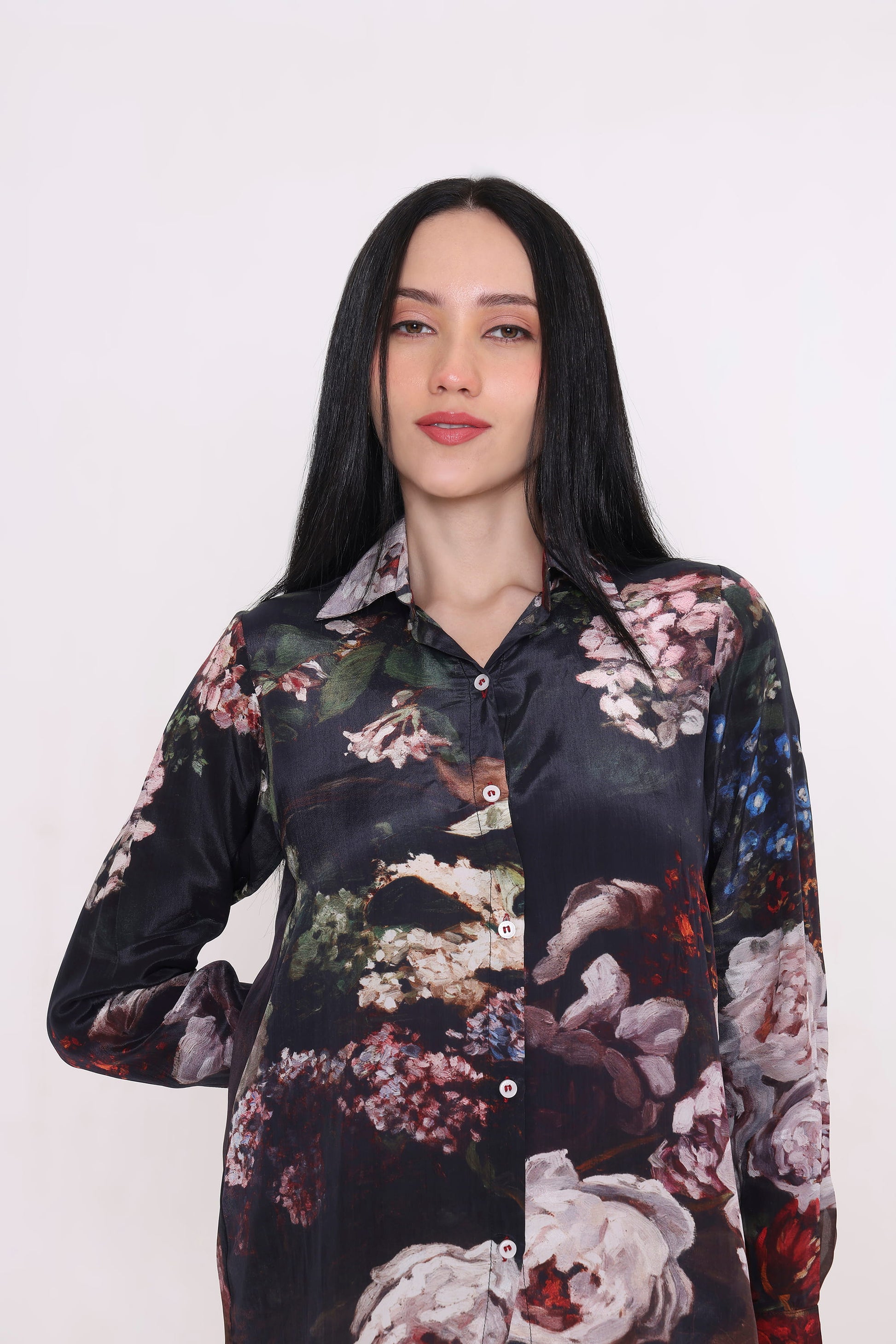 A woman with long black hair wearing a long-sleeve button-up shirt with a dark base and a large floral print in pink, white, green, and red, photographed against a plain light background.