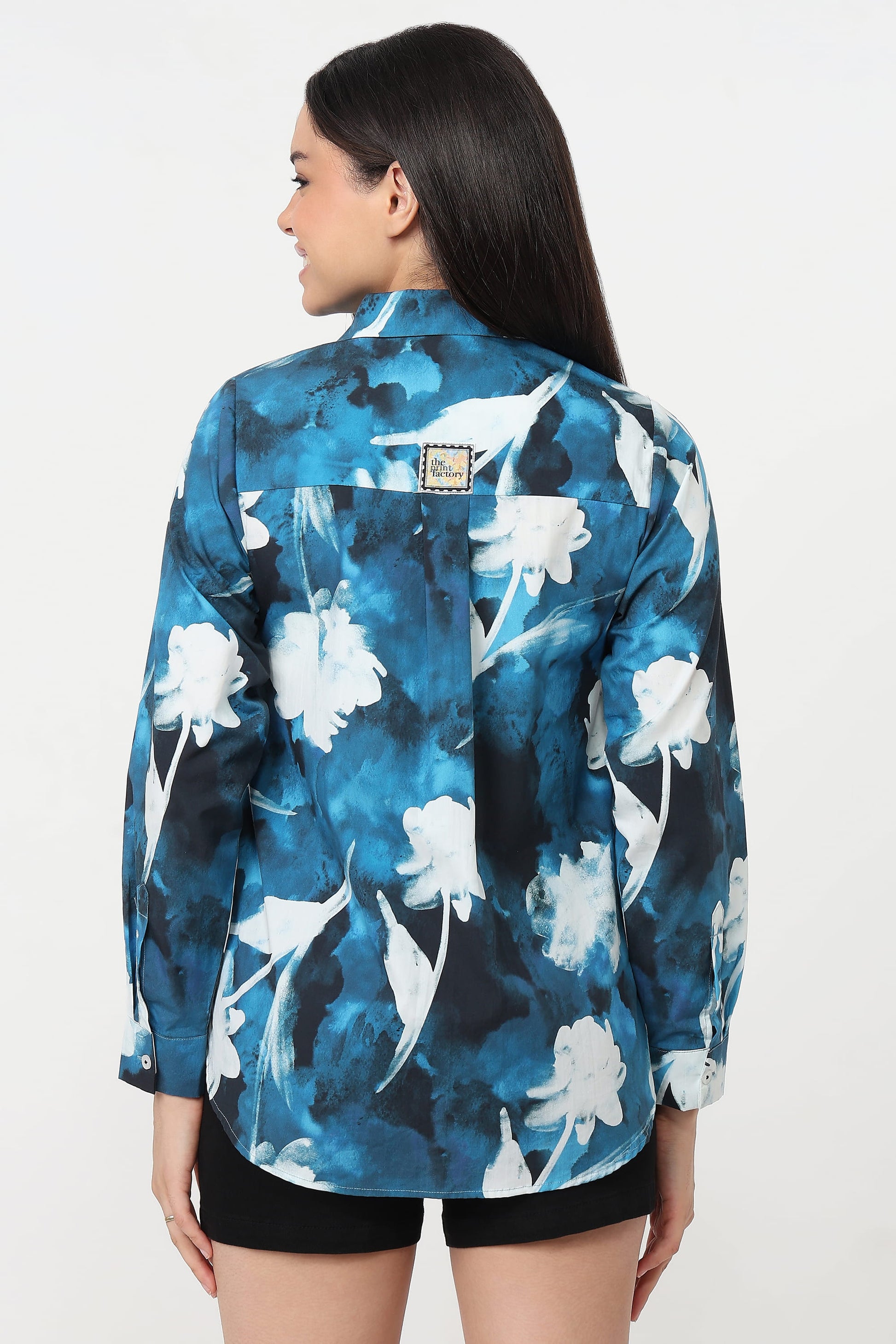 A woman shown from the back wearing a blue and white floral print button-up shirt with black shorts, photographed against a plain light background.