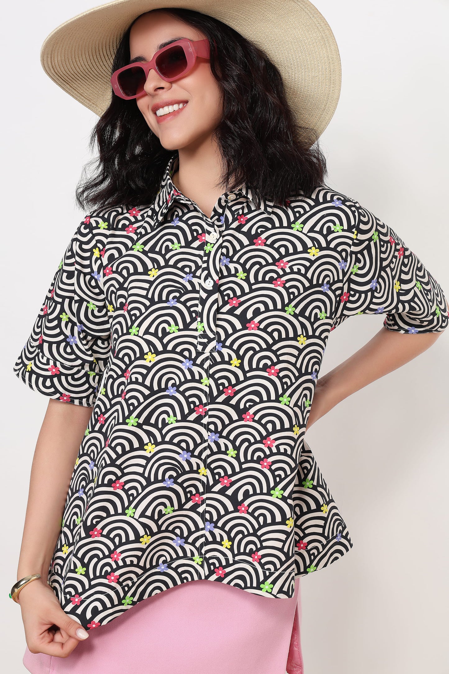 A woman wearing a short-sleeve button-up shirt with a black, white, and gray wave pattern accented by small colorful floral details, styled with a beige straw hat, pink sunglasses, and a pink skirt against a plain white background.