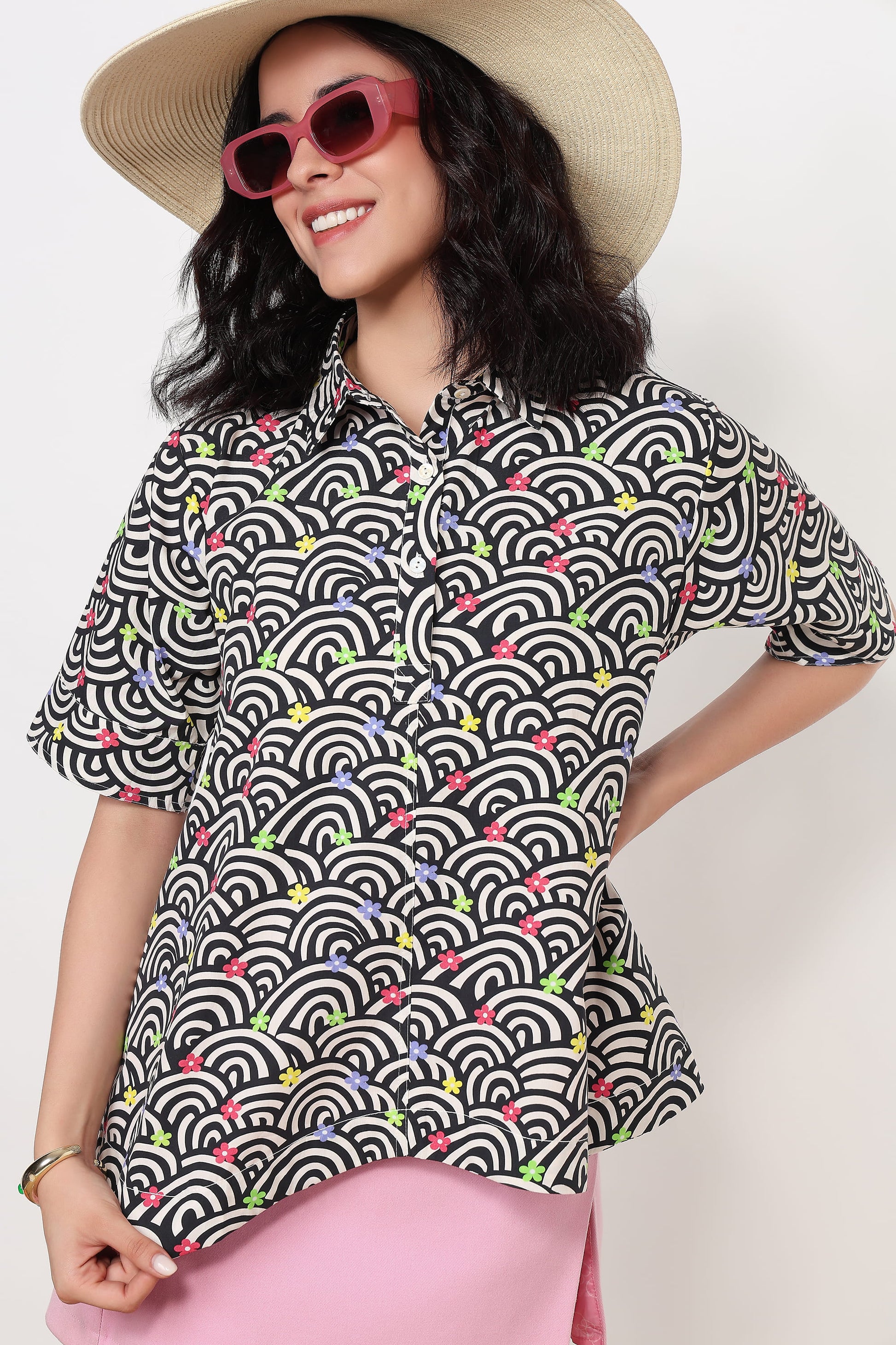 A woman wearing a short-sleeve button-up shirt with a black, white, and gray wave pattern accented by small colorful floral details, styled with a beige straw hat, pink sunglasses, and a pink skirt against a plain white background.