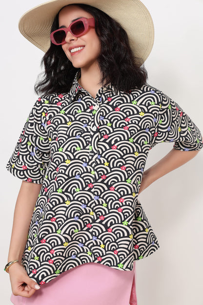 A woman wearing a short-sleeve button-up shirt with a black, white, and gray wave pattern accented by small colorful floral details, styled with a beige straw hat, pink sunglasses, and a pink skirt against a plain white background.