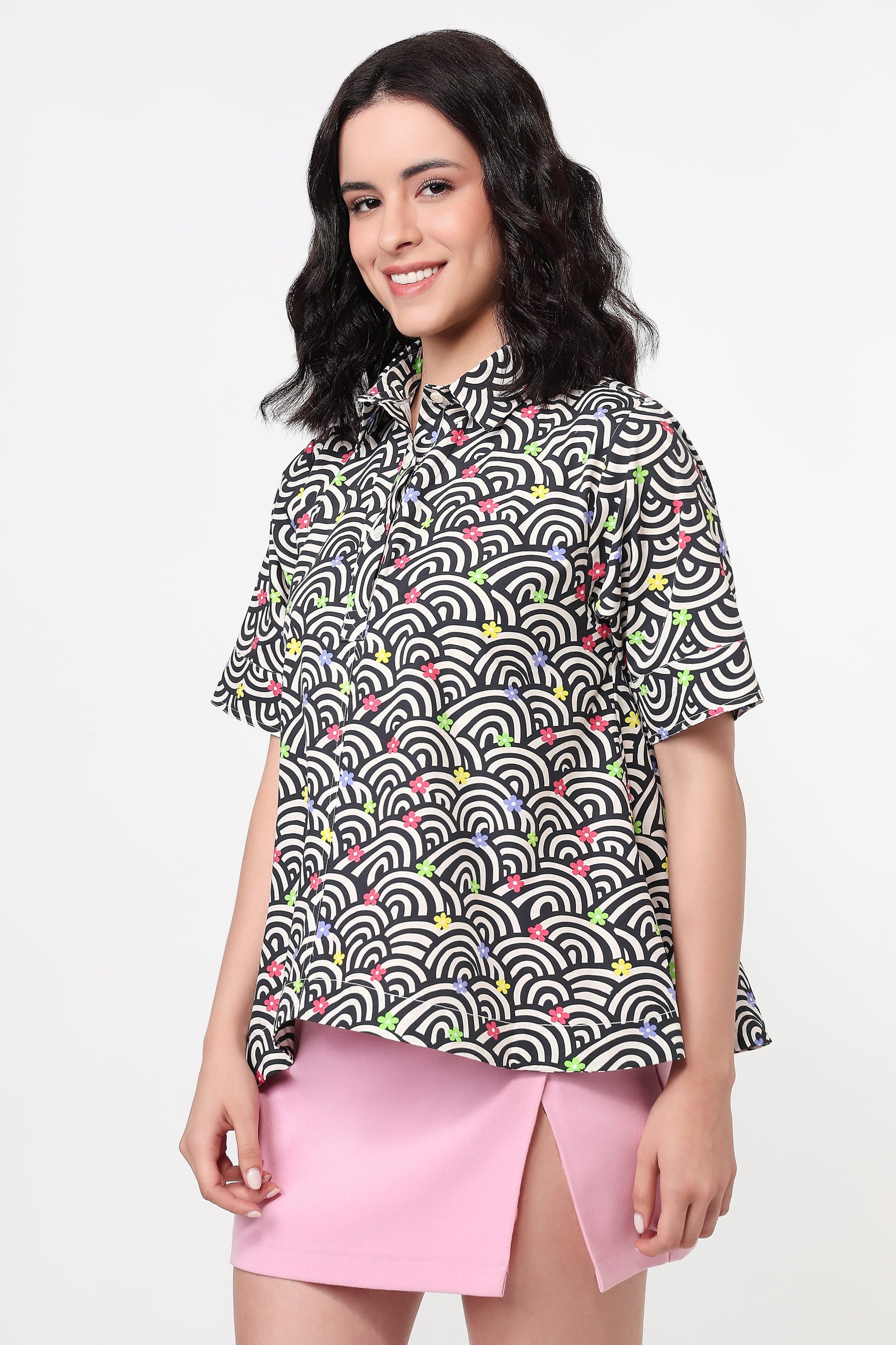 A woman modeling a short-sleeve button-up shirt with a black, white, and gray wave pattern accented with small pink and green details, paired with a pink skirt, against a plain white background.