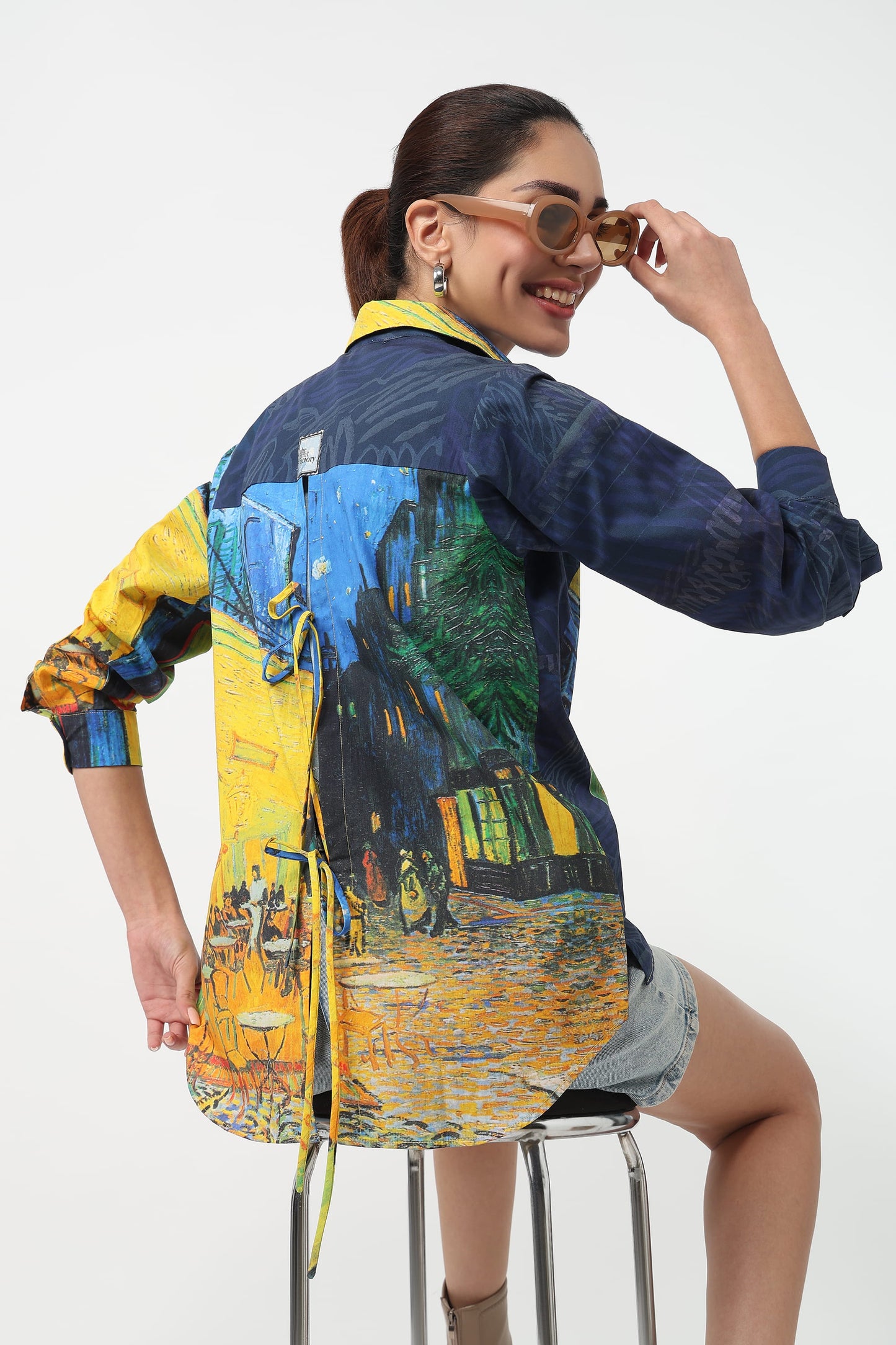 A woman wearing a blue denim shirt with a Van Gogh-inspired print in yellow, green, and blue, posing on a metal stool against a white background.