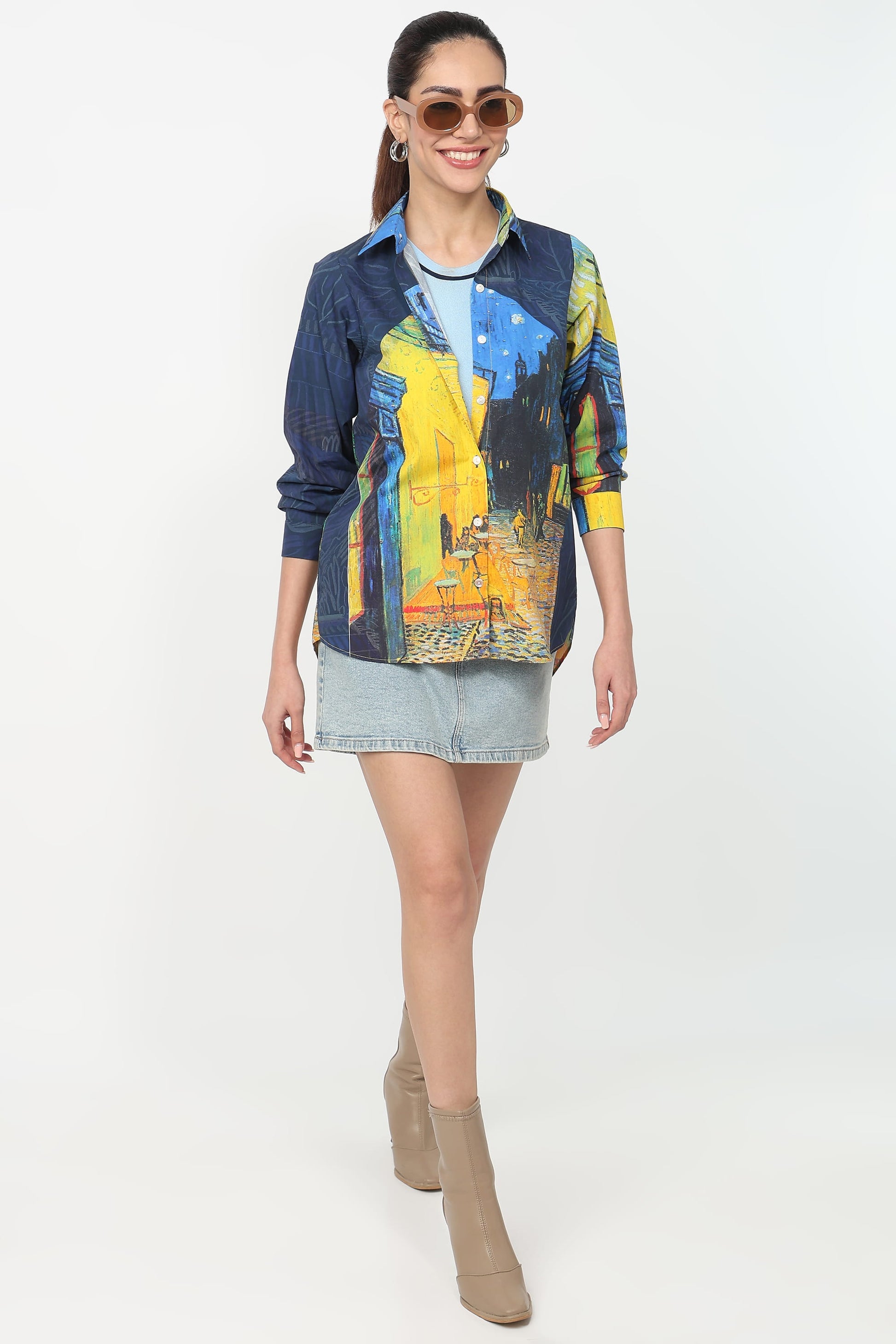 A woman modeling a long-sleeve button-up shirt with a blue and yellow abstract art print, paired with a denim mini skirt and beige ankle boots, against a plain white studio background.