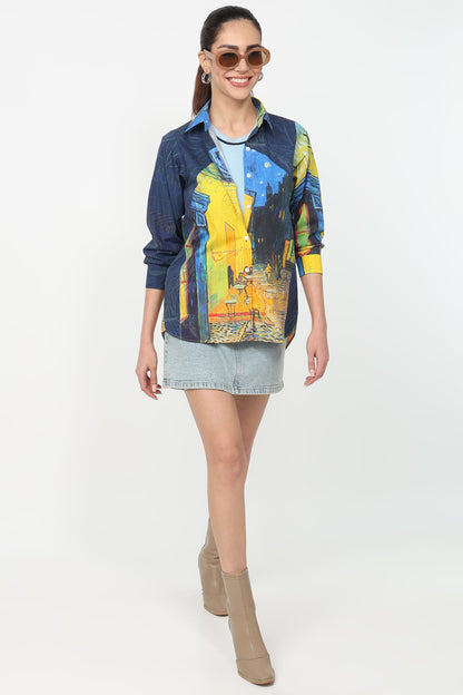 A woman modeling a long-sleeve button-up shirt with a blue and yellow abstract art print, paired with a denim mini skirt and beige ankle boots, against a plain white studio background.
