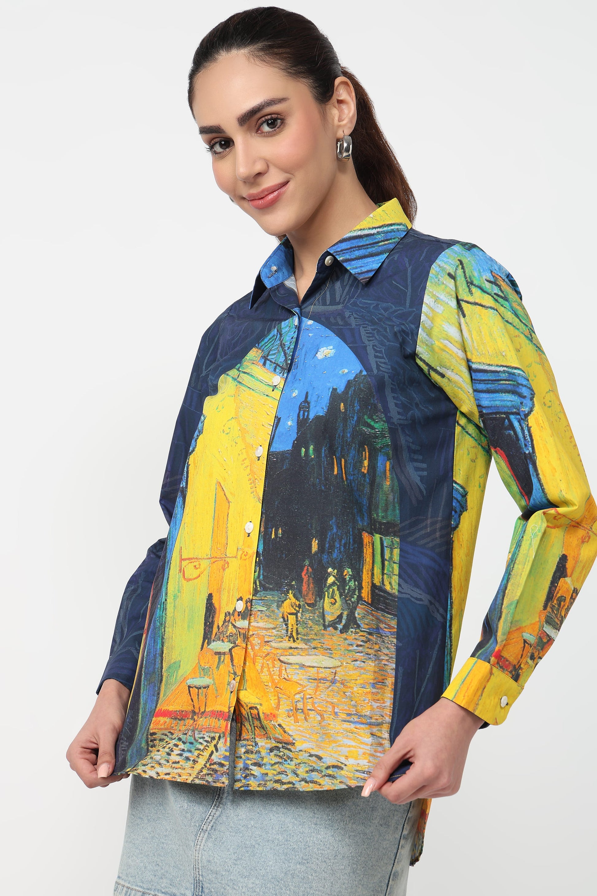 A woman wearing a long-sleeve button-up shirt with a Van Gogh-inspired street scene print in yellow, blue, and dark navy tones, paired with light gray jeans against a plain light gray studio background.