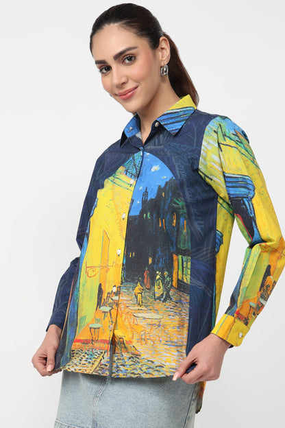 A woman wearing a long-sleeve button-up shirt with a Van Gogh-inspired street scene print in yellow, blue, and dark navy tones, paired with light gray jeans against a plain light gray studio background.