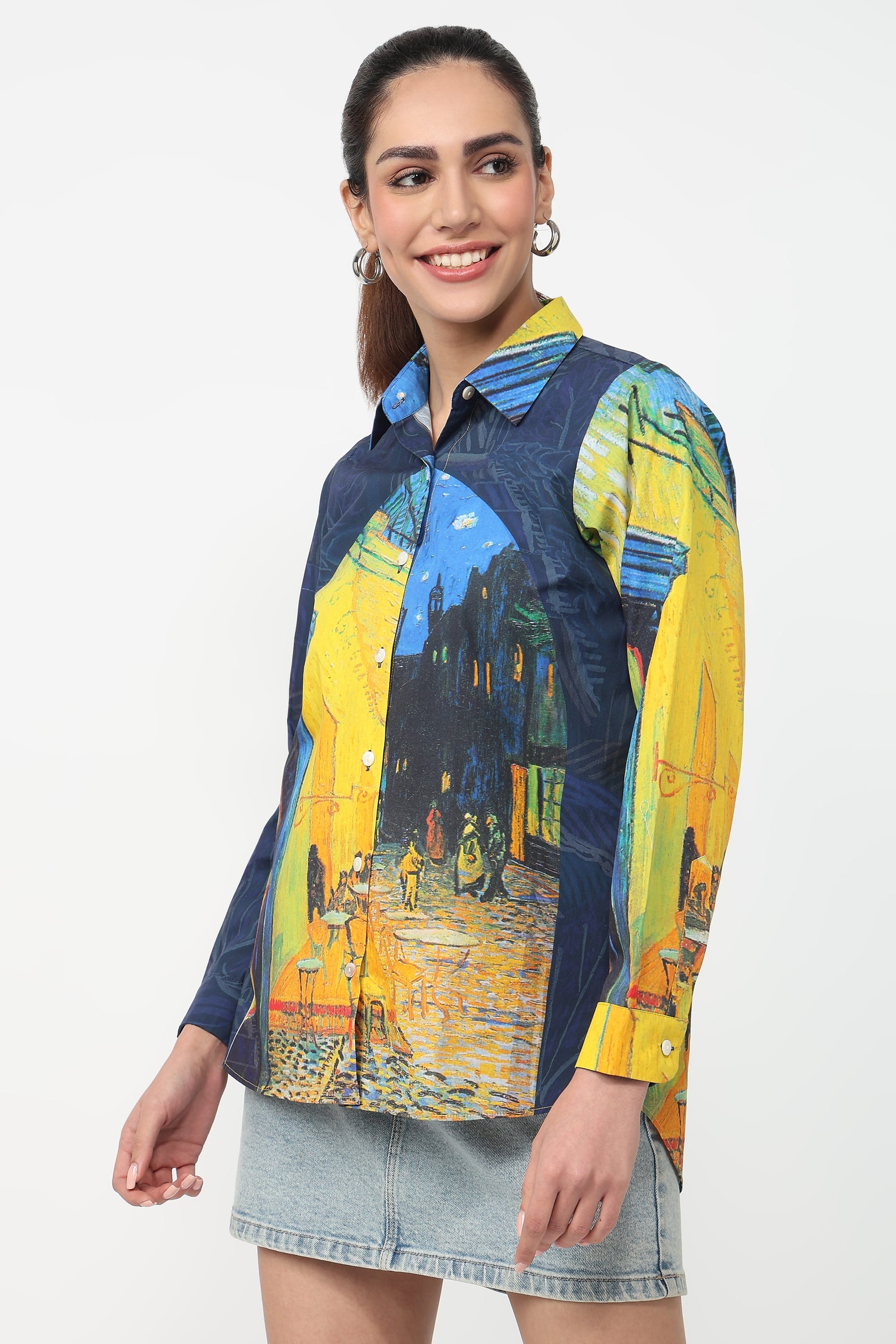 A woman modeling a long-sleeve button-up shirt with a Van Gogh-inspired print featuring blue denim-style areas and bright yellow, orange, and blue artwork panels, paired with a light denim skirt against a plain white background.