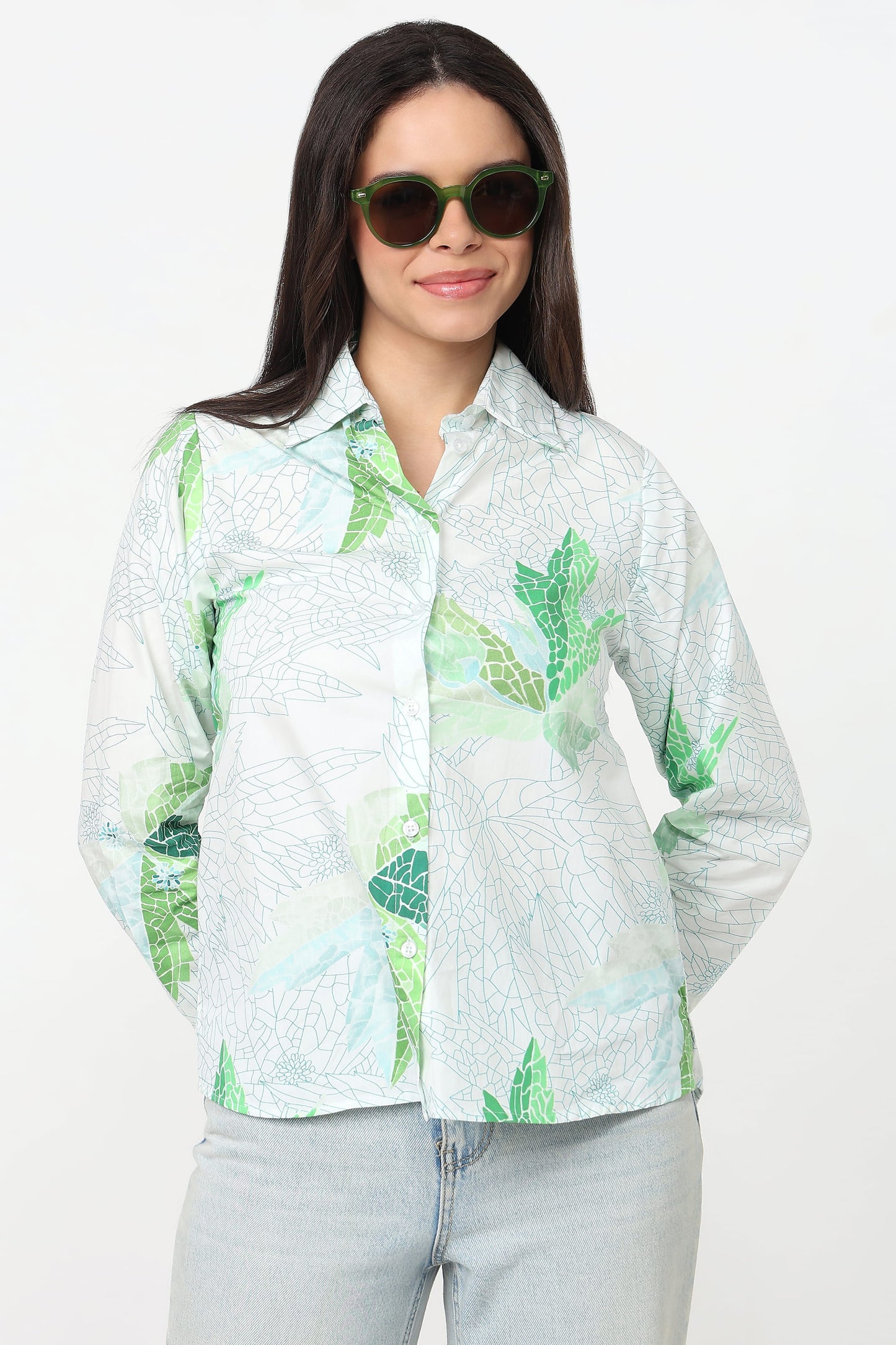 A woman wearing a white long-sleeve button-up shirt with a green tropical leaf pattern, paired with light-wash jeans and sunglasses, against a plain white background.