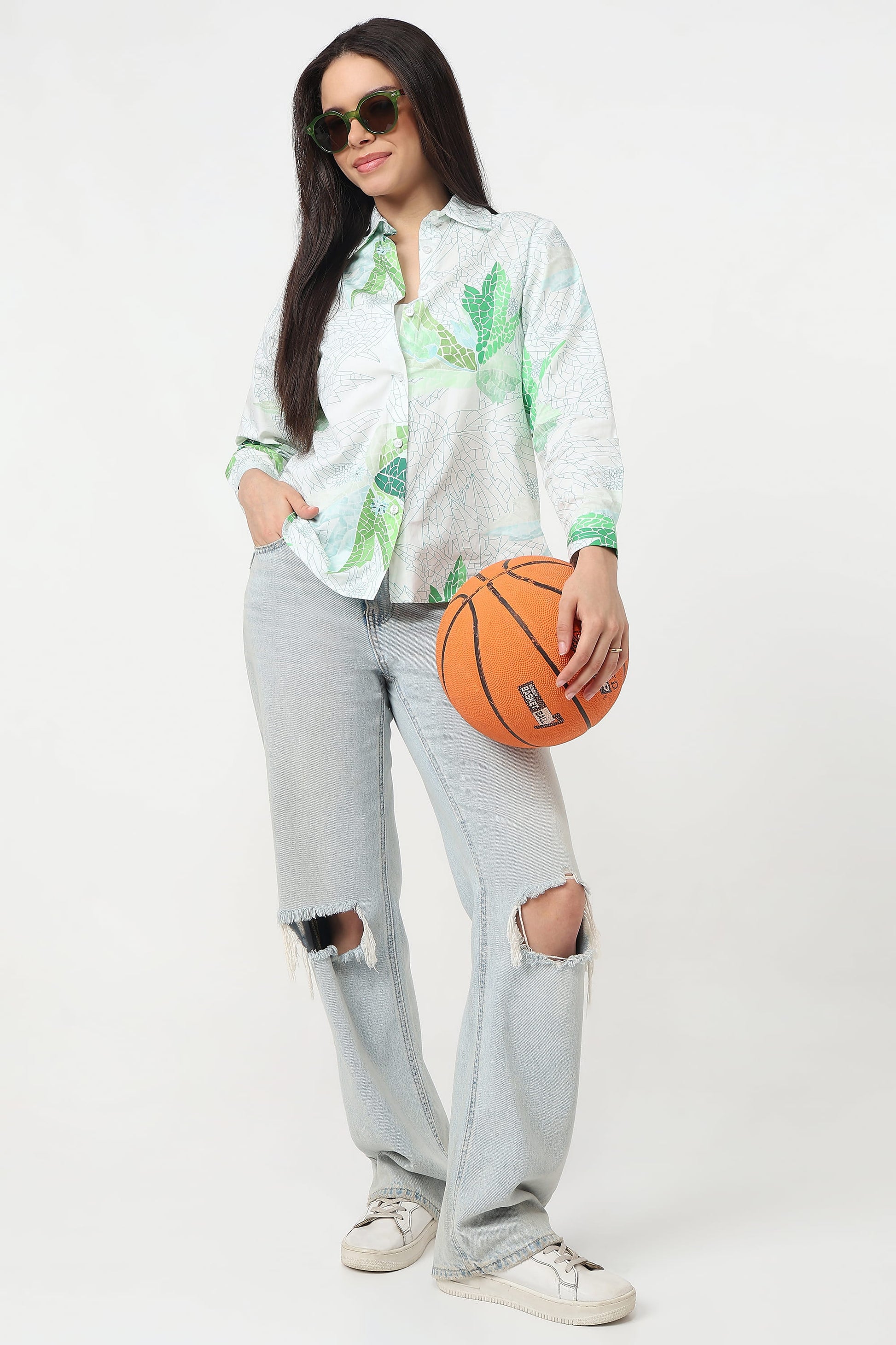 A woman wearing a white button-up shirt with a green tropical leaf print, paired with light-wash ripped jeans, white sneakers, and sunglasses, holding an orange basketball against a plain white studio background.