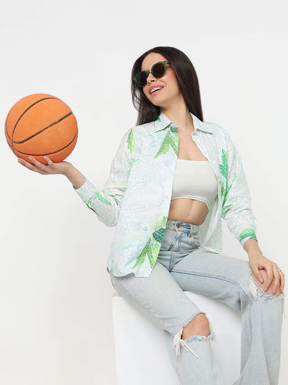 A woman wearing a white button-up shirt with a green palm leaf pattern, light-wash ripped jeans, and sunglasses, holding an orange basketball while sitting on a white block against a white background.