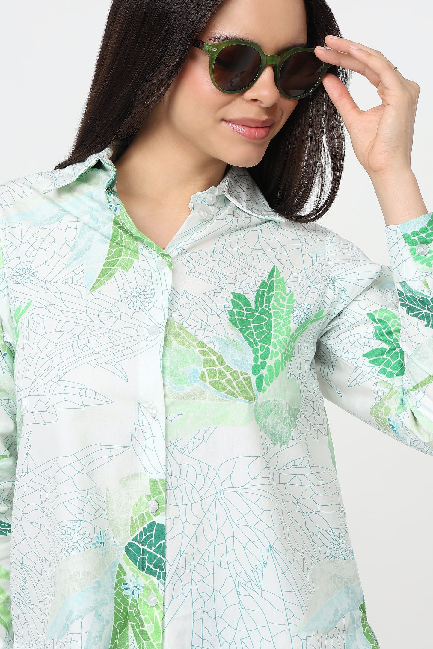 A woman wearing a white button-up shirt with a green and light blue leaf pattern, styled with green-framed sunglasses against a plain white background.