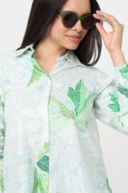 A woman wearing a white button-up shirt with a green and light blue leaf pattern, styled with green-framed sunglasses against a plain white background.