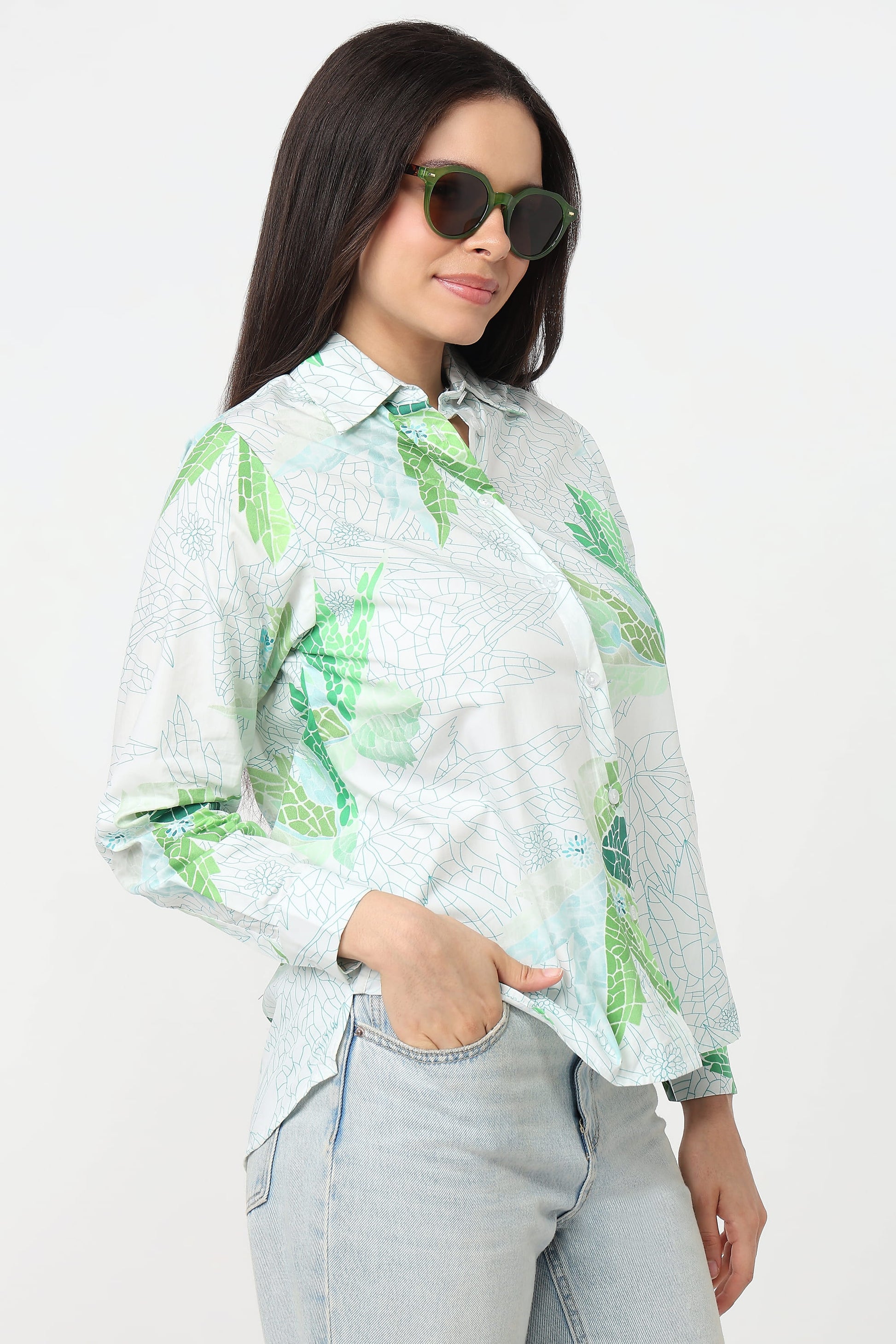 A woman wearing a light green and white tropical leaf print button-up shirt and light-wash jeans, posed against a plain white studio background.
