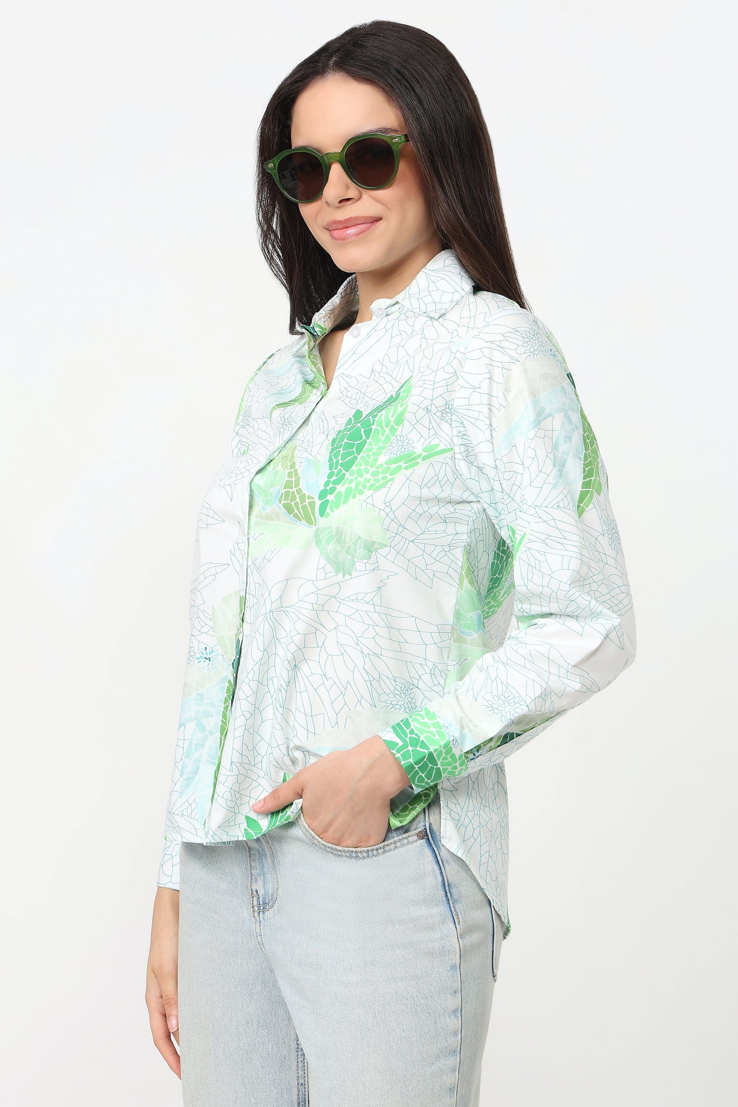 A woman wearing a white long-sleeve button-up shirt with a green tropical leaf pattern, paired with light-wash jeans and green-tinted sunglasses, against a plain white background.