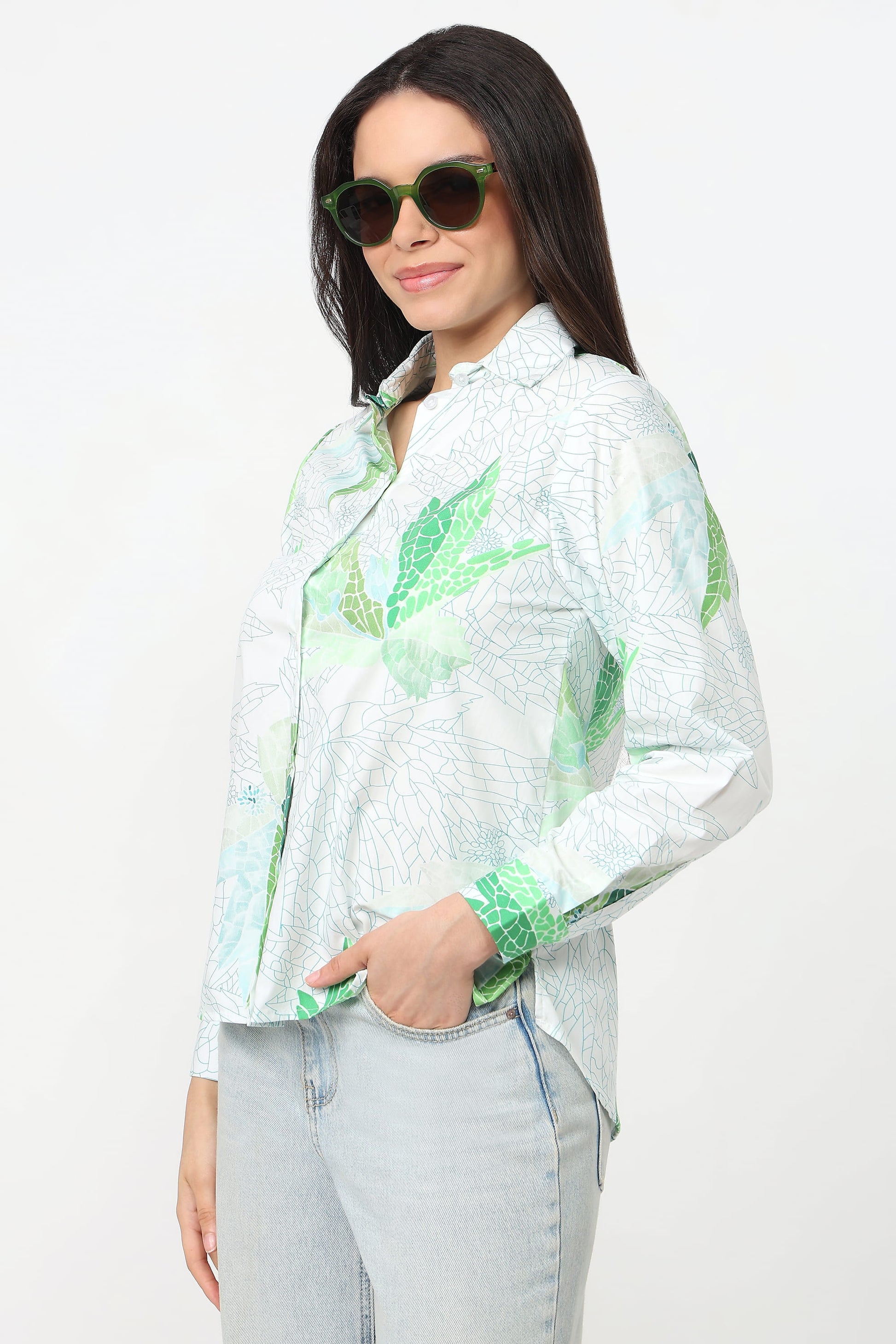 A woman wearing a white long-sleeve button-up shirt with a green tropical leaf pattern, paired with light-wash jeans and green-tinted sunglasses, against a plain white background.