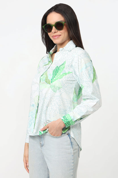 A woman wearing a white long-sleeve button-up shirt with a green tropical leaf pattern, paired with light-wash jeans and green-tinted sunglasses, against a plain white background.