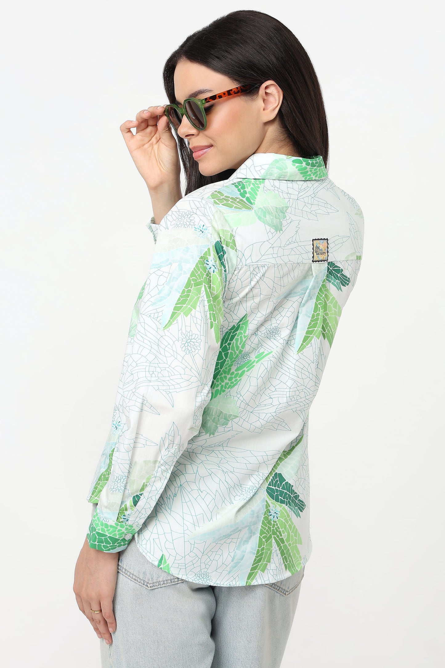 A woman wearing a light green and white tropical leaf print button-up shirt, styled with light gray jeans and tortoiseshell sunglasses, photographed against a plain white studio background.