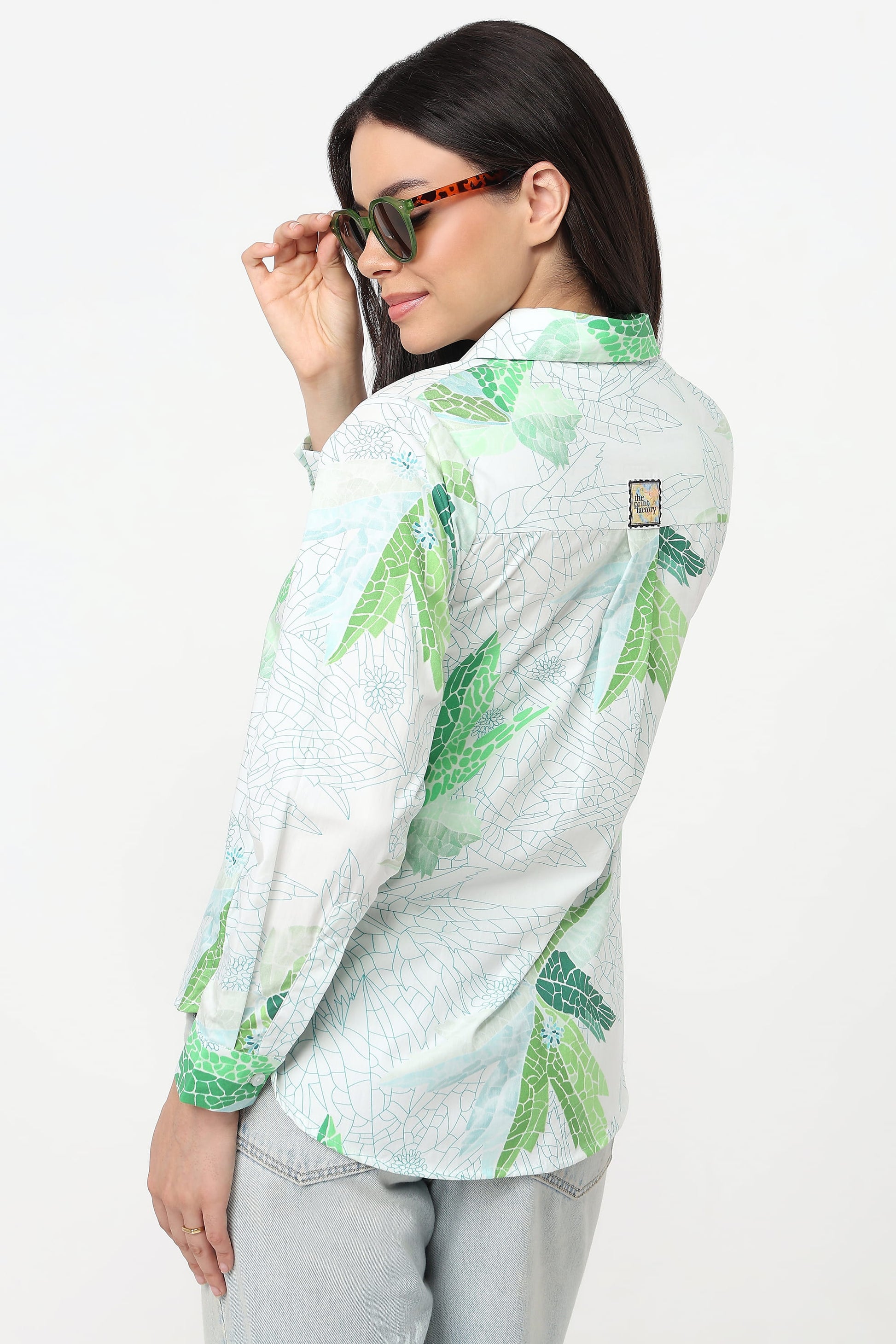 A woman wearing a light green and white tropical leaf print button-up shirt, styled with light gray jeans and tortoiseshell sunglasses, photographed against a plain white studio background.
