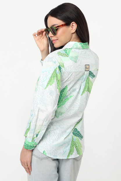 A woman wearing a light green and white tropical leaf print button-up shirt, styled with light gray jeans and tortoiseshell sunglasses, photographed against a plain white studio background.