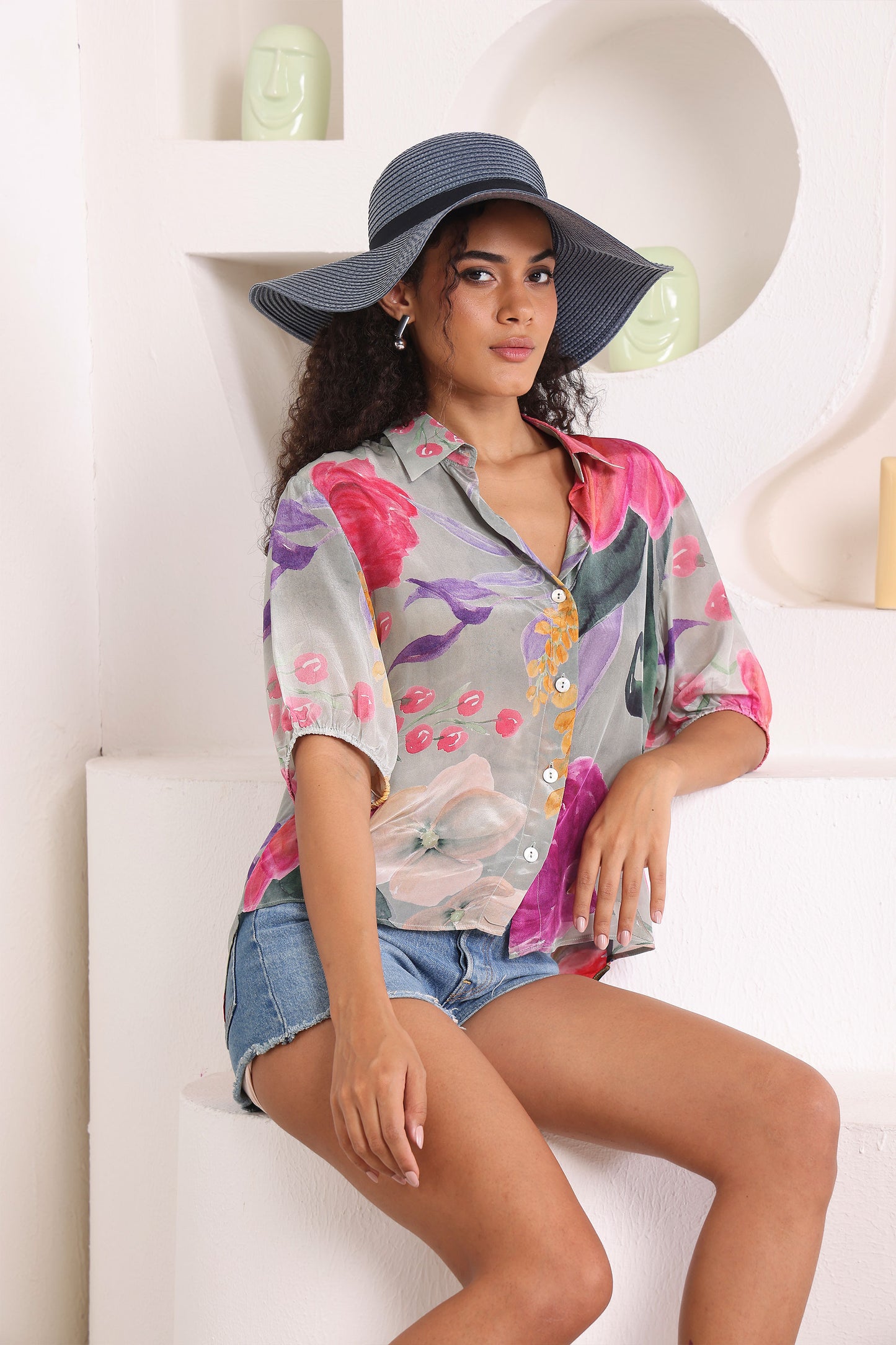 Woman wearing a colorful shirt and denim shorts sitting on a white surface.