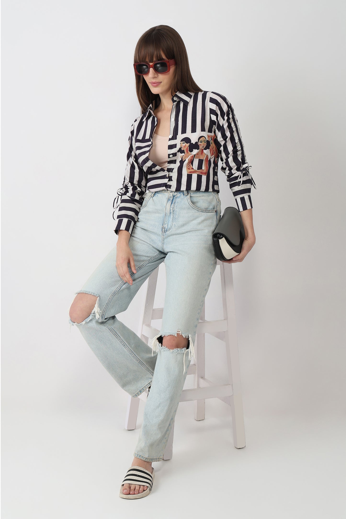 Woman wearing a black and white striped shirt with a graphic design, light blue jeans, and sandals on a white background.