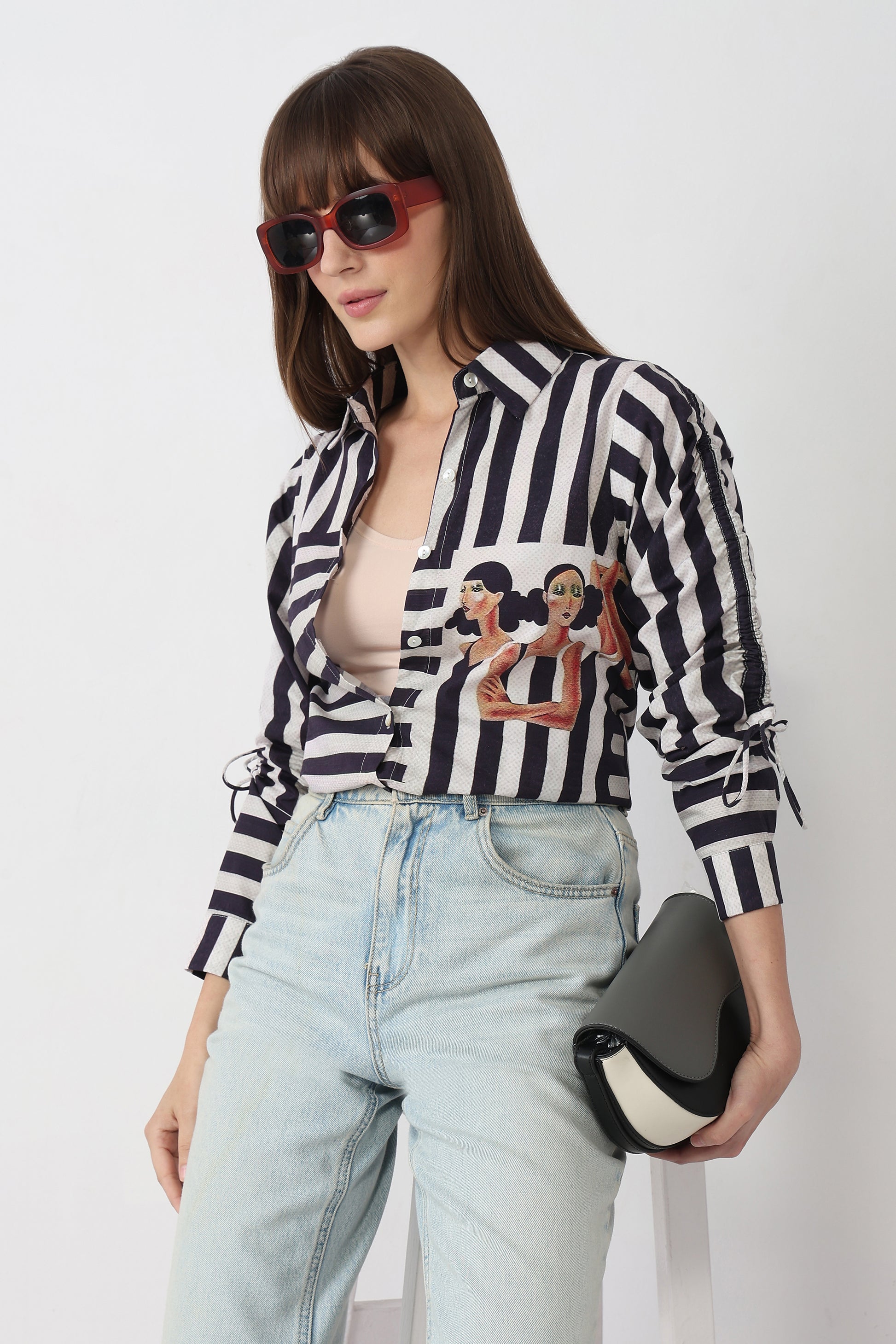 Woman wearing a black and white striped shirt with graphic design, light blue jeans, and sunglasses on a white background.