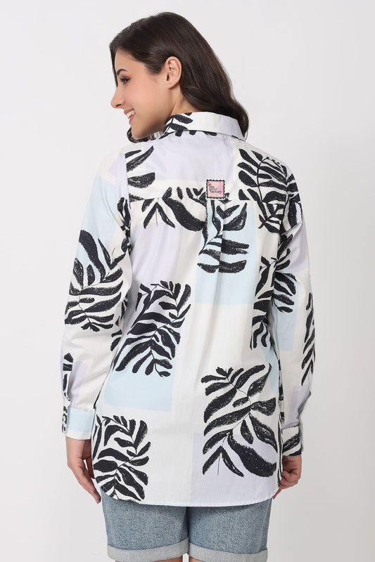 A woman shown from the back wearing a white long-sleeve button-up shirt with a black tropical leaf print and light blue color blocks, paired with denim shorts against a plain light gray background.