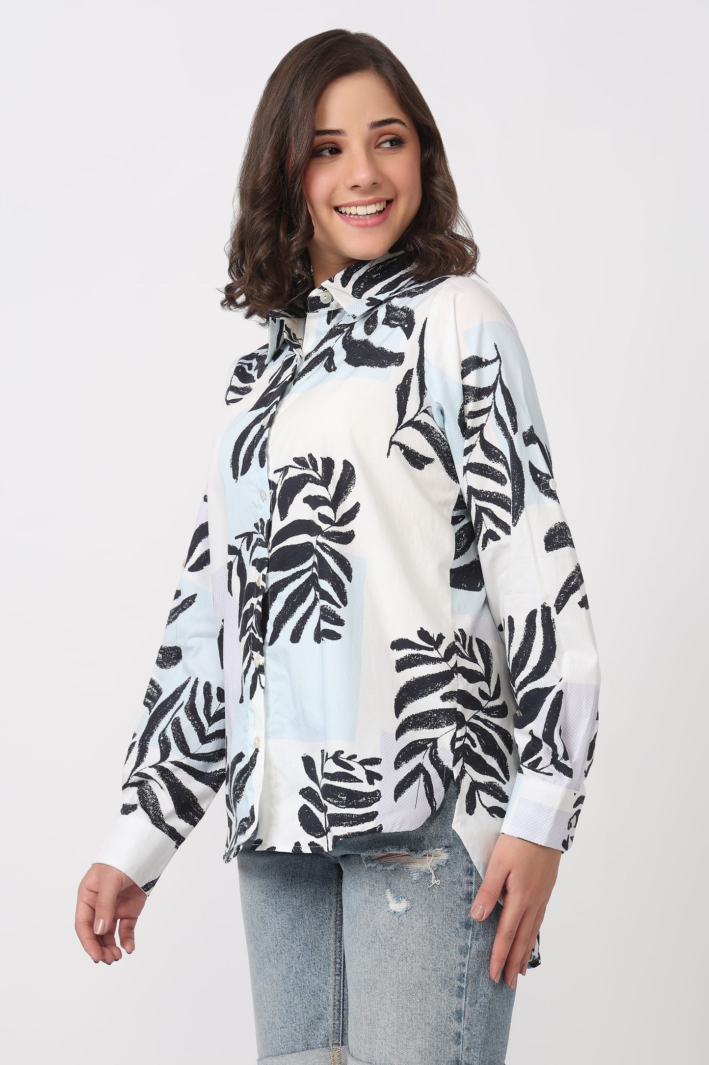 A woman wearing a white long-sleeve blouse with a black tropical leaf print, paired with light-wash distressed jeans, against a light gray studio background.