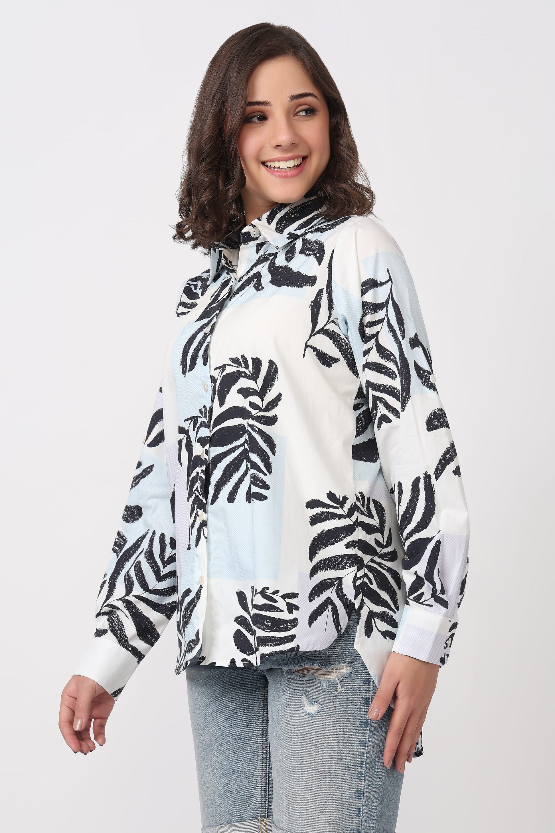A woman wearing a white long-sleeve blouse with a black tropical leaf print, paired with light-wash distressed jeans, against a light gray studio background.