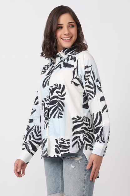 A woman wearing a white long-sleeve blouse with a black tropical leaf print, paired with light-wash distressed jeans, against a light gray studio background.
