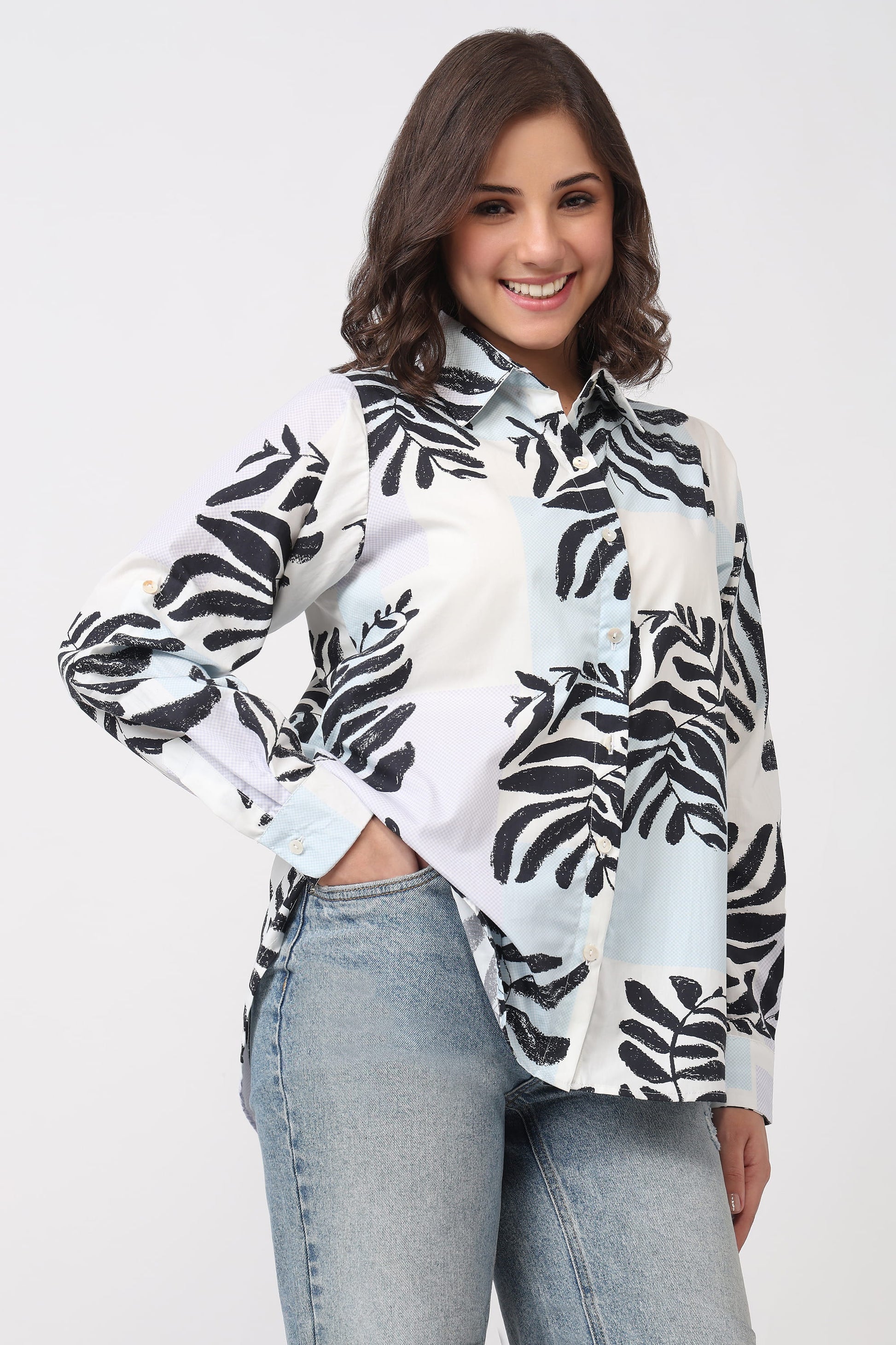 A woman wearing a white button-up shirt with a black tropical leaf print, paired with light-wash blue jeans, against a plain light gray background.