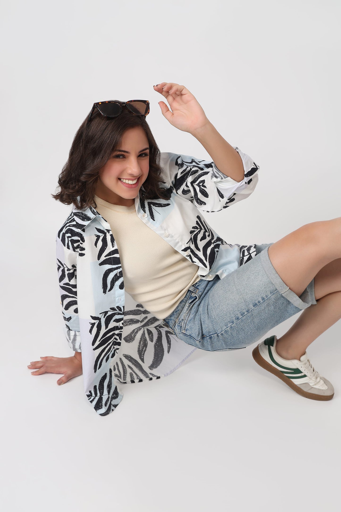 A woman sitting on a light gray studio floor wearing a white button-up shirt with a black palm leaf pattern, a beige ribbed top, light-wash denim shorts, and white sneakers with green stripes.