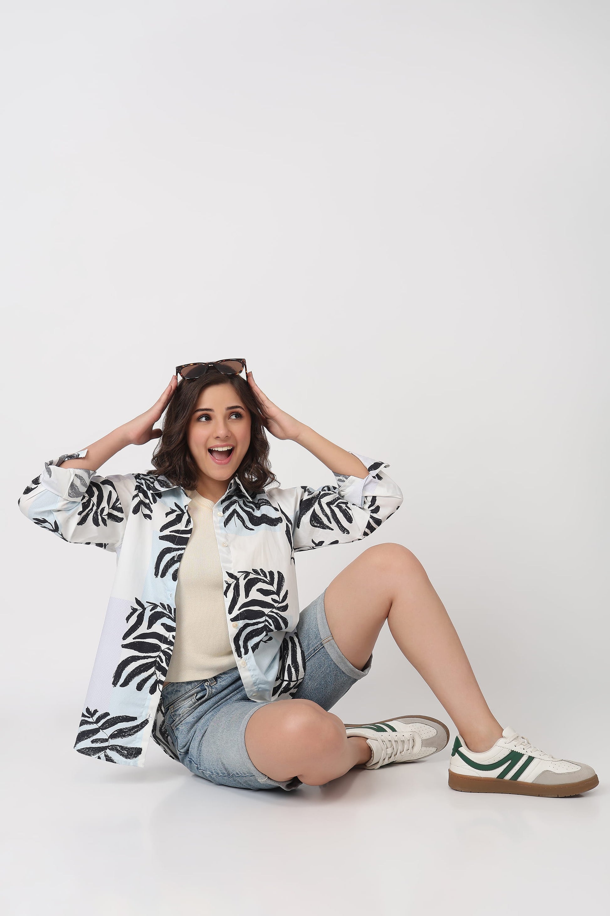 A woman sitting on a white studio floor wearing a white button-up shirt with a black palm leaf pattern, denim shorts, and white sneakers with green stripes.