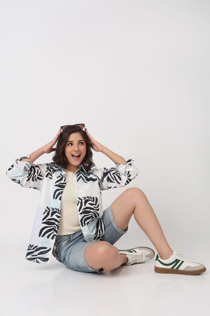 A woman sitting on a white studio floor wearing a white button-up shirt with a black palm leaf pattern, denim shorts, and white sneakers with green stripes.