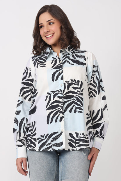 A woman wearing a white button-up shirt with a black tropical leaf print, paired with light-wash jeans, against a plain light gray studio background.