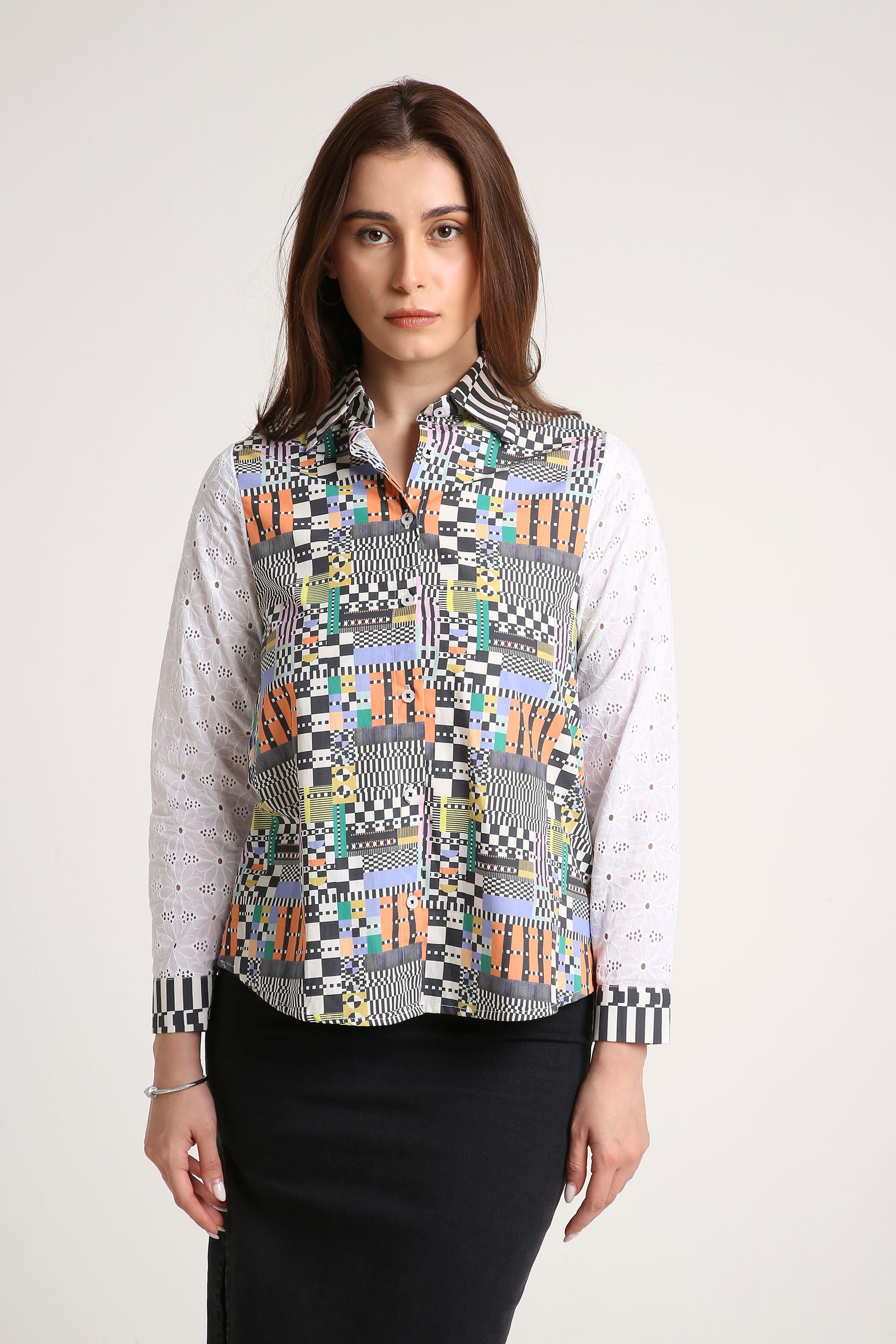 A woman wearing a long-sleeve button-up shirt with a colourful geometric patchwork pattern on the front and white eyelet sleeves, paired with a black skirt, against a light grey studio background.