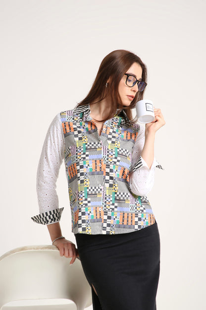 A woman wearing a long-sleeve button-up shirt with a colorful geometric patchwork pattern over a white base, paired with a black skirt, holding a white mug against a plain light background.