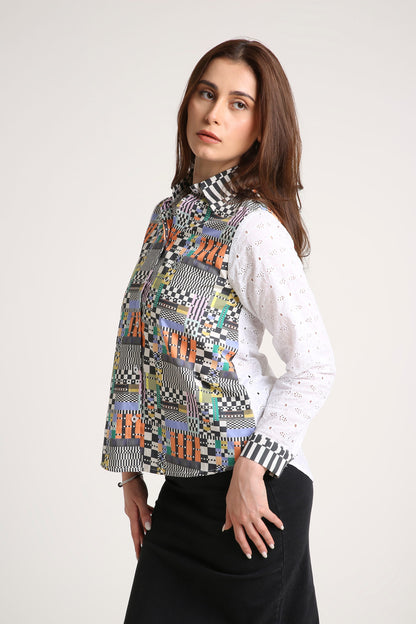 A woman wearing a long-sleeve button-up shirt with a colourful geometric patchwork pattern on the front and white eyelet sleeves, paired with a black skirt, against a light grey studio background.