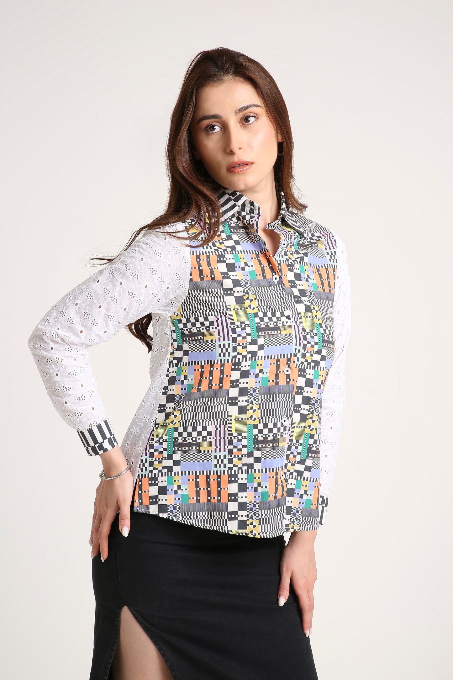 A woman wearing a long-sleeve button-up shirt with a colourful geometric patchwork pattern on the front and white eyelet sleeves, paired with a black skirt, against a light grey studio background.