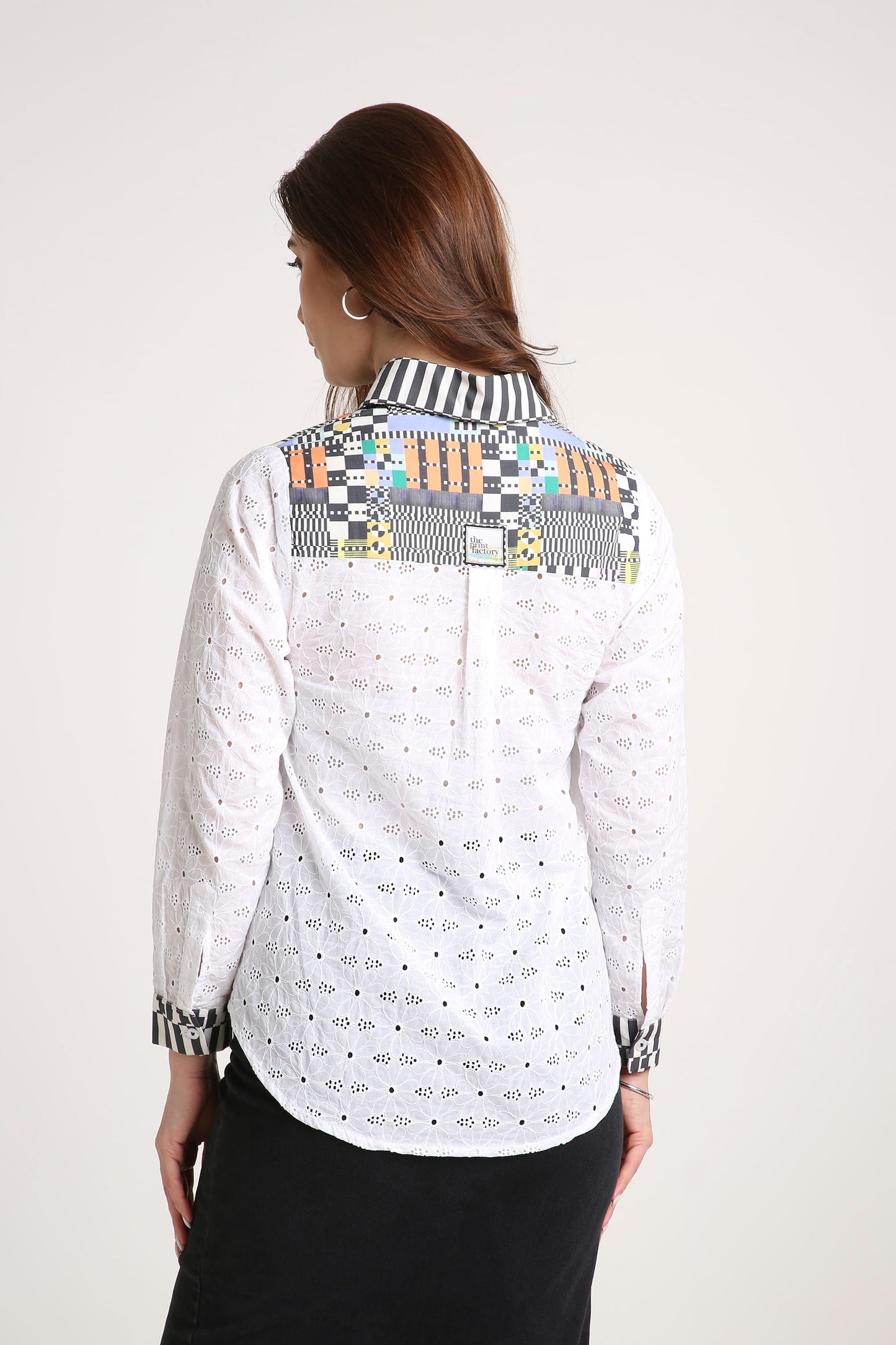 A woman shown from the back wearing a white long-sleeve eyelet blouse with a multicolour geometric patchwork design across the shoulders and black skirt, set against a plain light grey studio background.