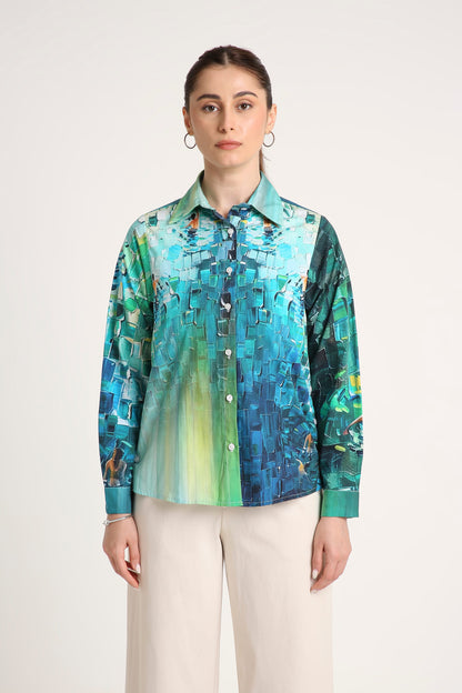 A woman wearing a long-sleeve button-up shirt with an abstract geometric print in shades of teal, blue, green, and yellow, paired with light beige trousers against a plain light background.